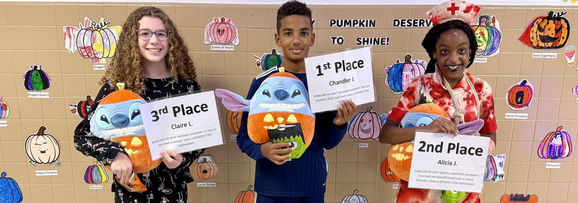 Three children holding plush toys and award signs, smiling in front of a decorated wall.