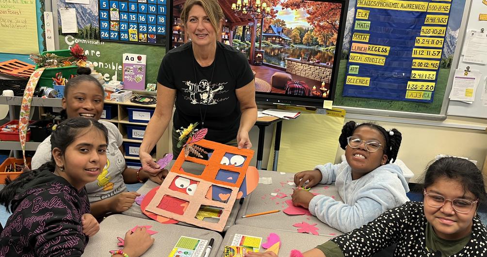 Teacher assisting students at a table with art projects and colorful materials.