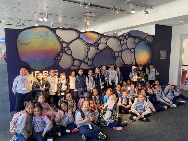 Class of students gathered in front of a colorful wall mural, celebrating their visit.