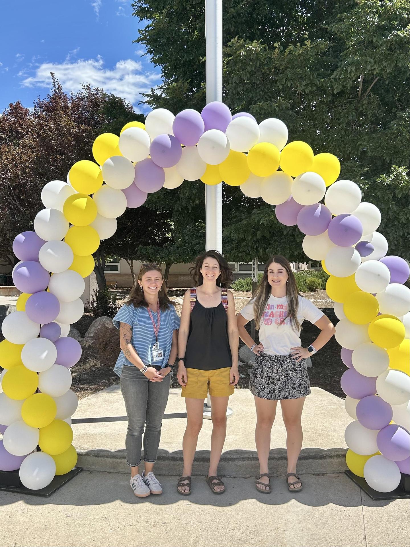 Three female teachers standing under a balloon arch with a tree in the background
