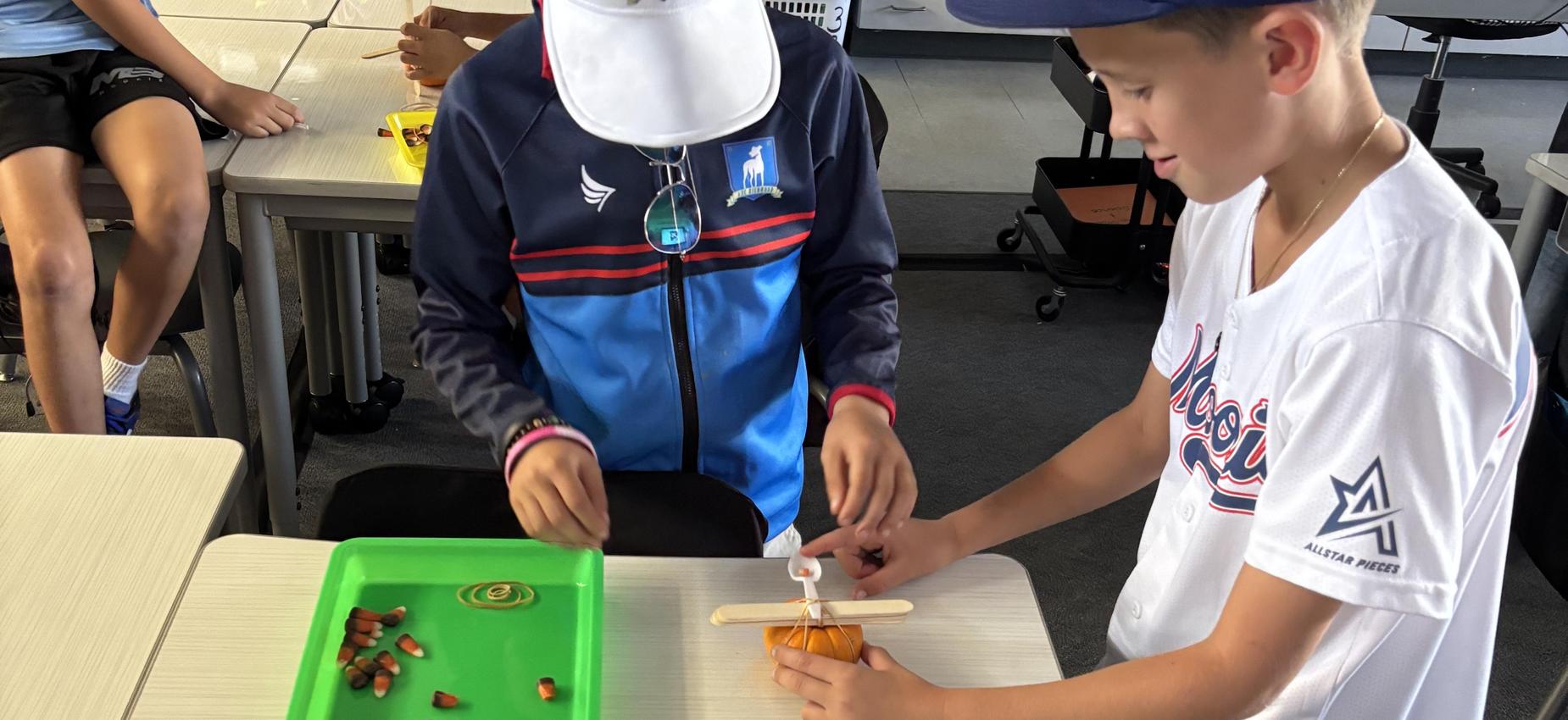 Two students constructing a small project with materials and a pumpkin base.