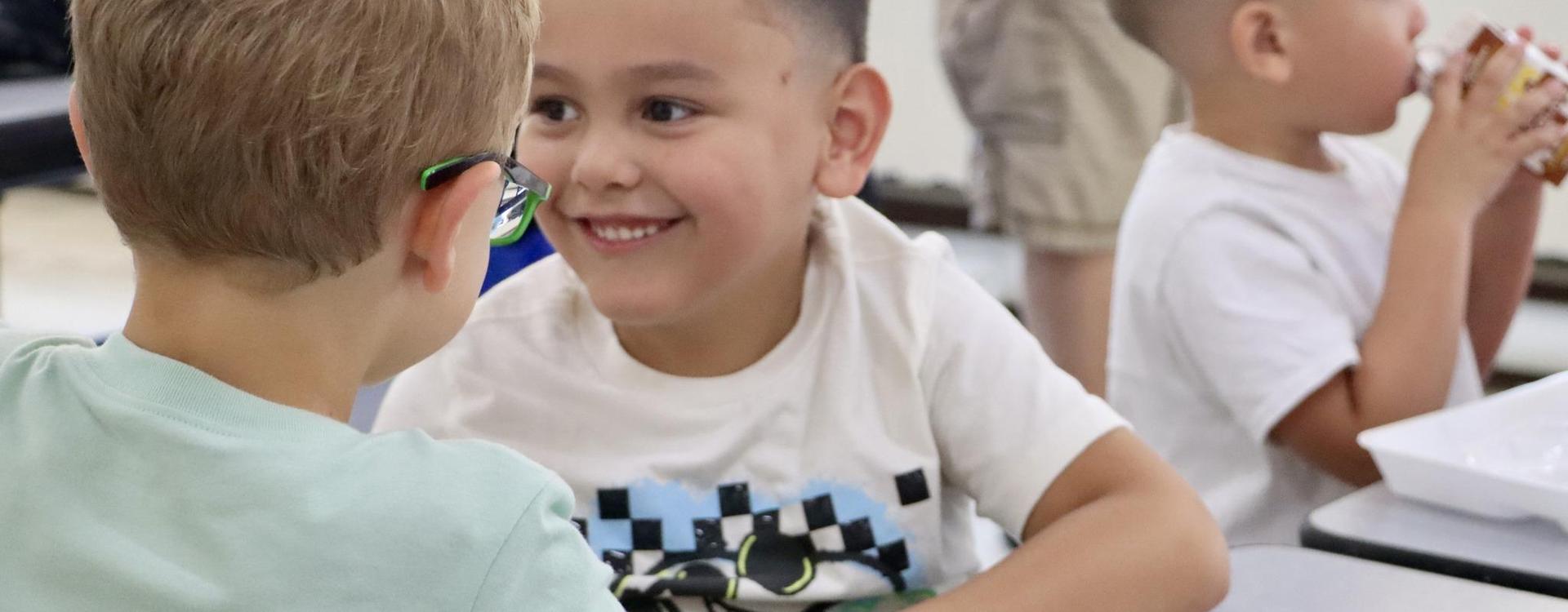 Two boys laughing and talking at a cafeteria table, with other children nearby.