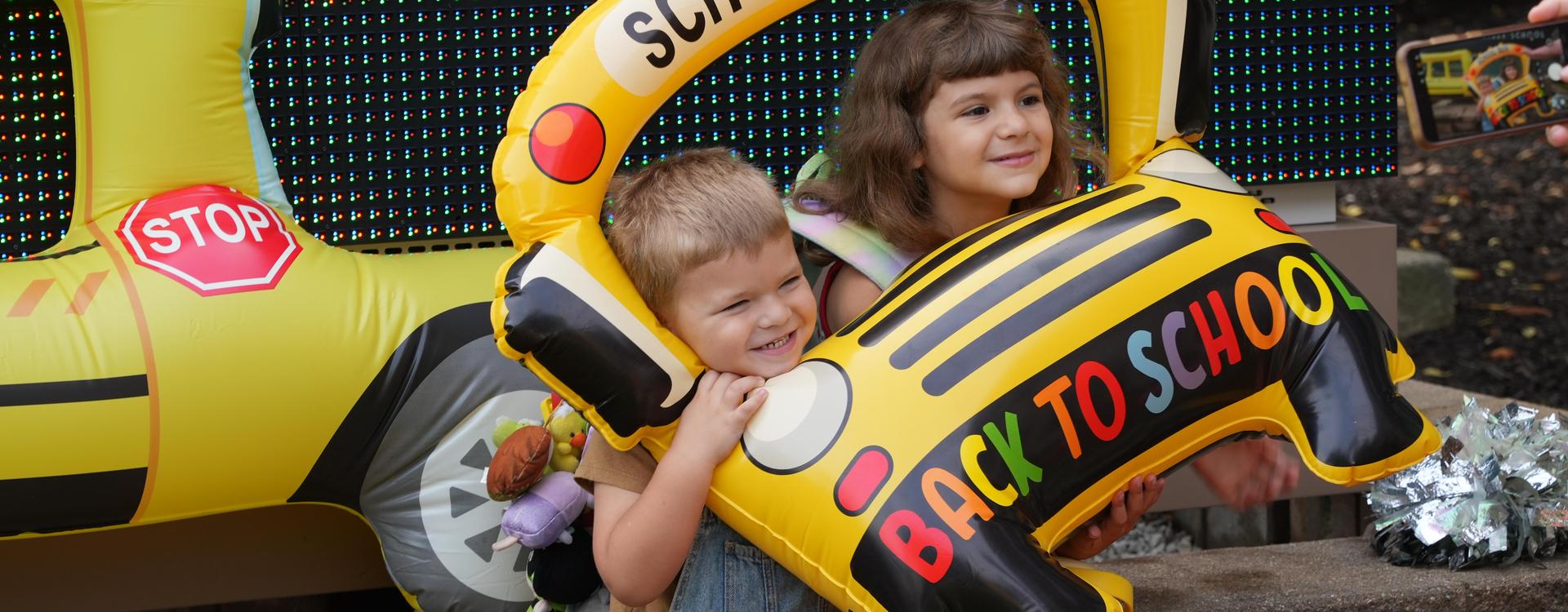Child holding a yellow inflatable school bus with 'BACK TO SCHOOL' text, smiling beside a girl.