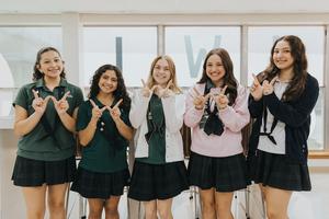 Five students in uniforms stand together indoors, making 'W' hand signs with smiles.