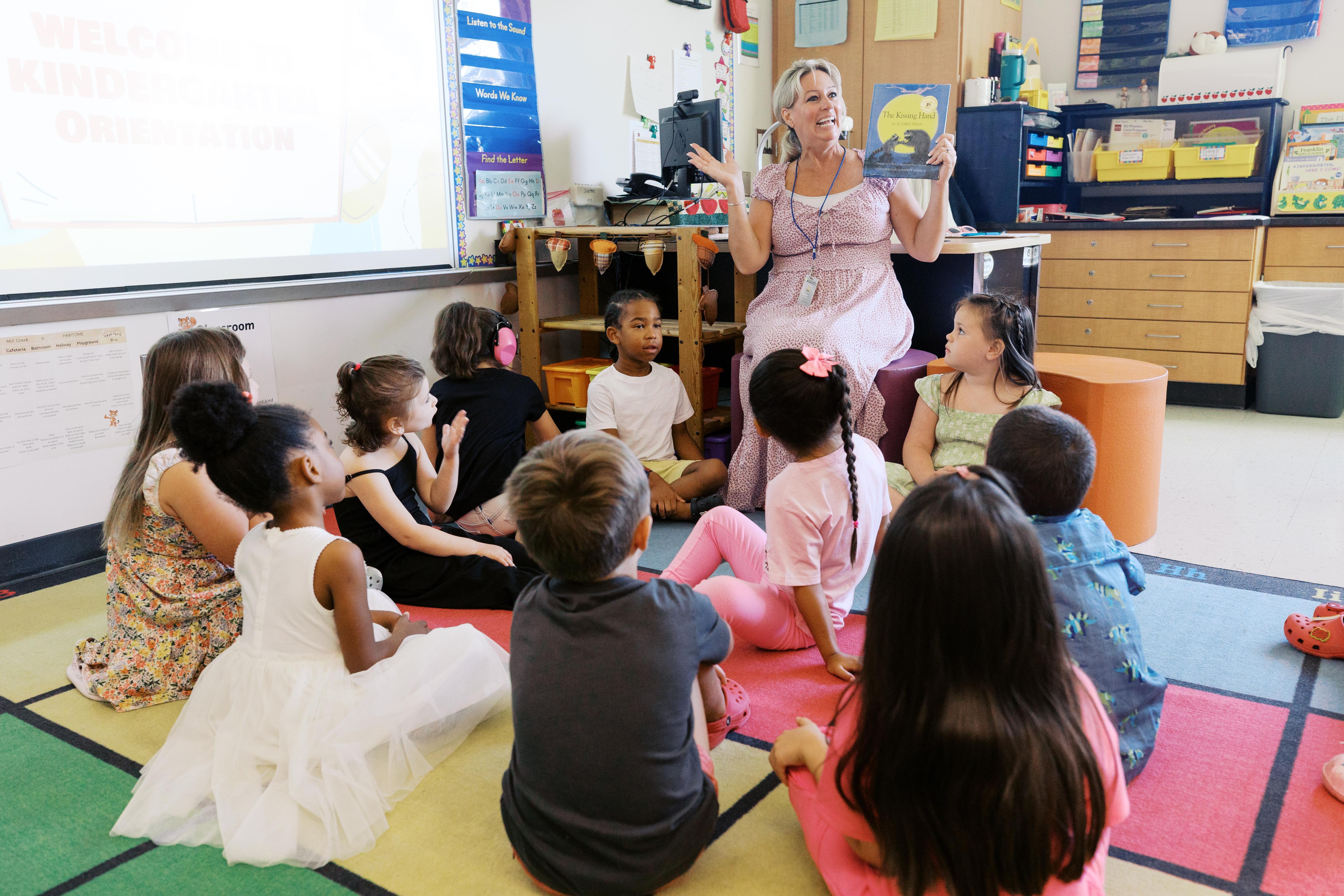 A teacher reads a book to students, who sit cross legged on the floor.