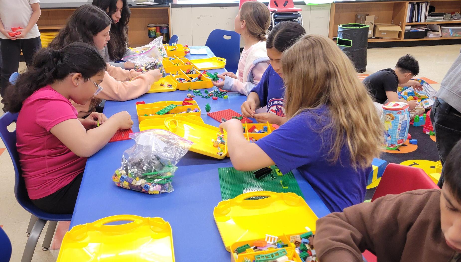 Group of children building with LEGO on a large table, surrounded by various pieces.