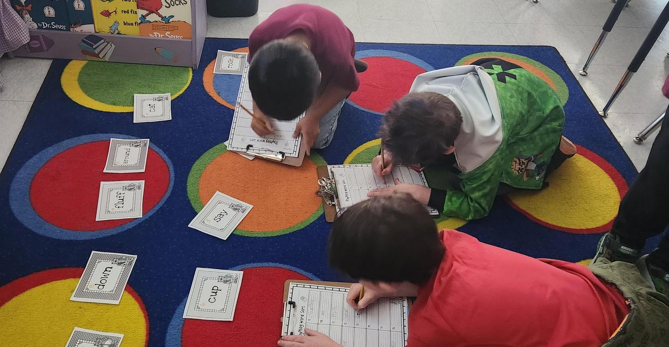 Three children writing on clipboards on a colorful carpet with cards around them.