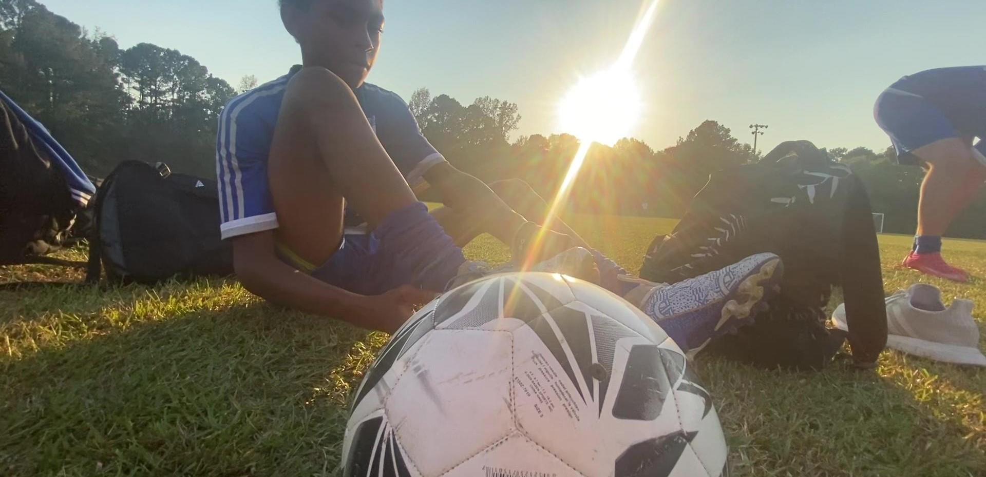 Soccer player tying cleats with a ball in foreground and sunset in the background.