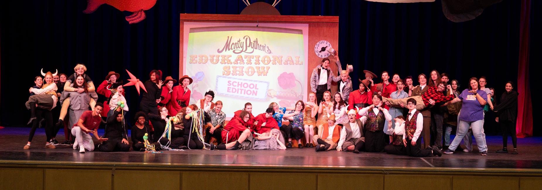 A large group of performers in costumes poses on stage for Monty Pythons Edukational Show