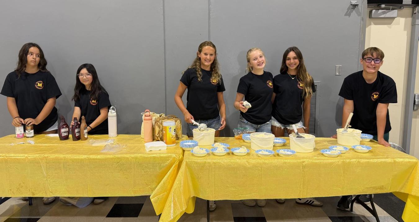 Six young people preparing food at a table with condiments and toppings.