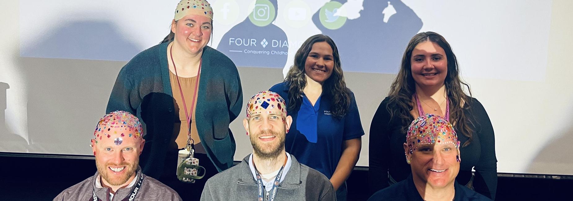 Group of five individuals posing with colorful hats in front of a projector screen.