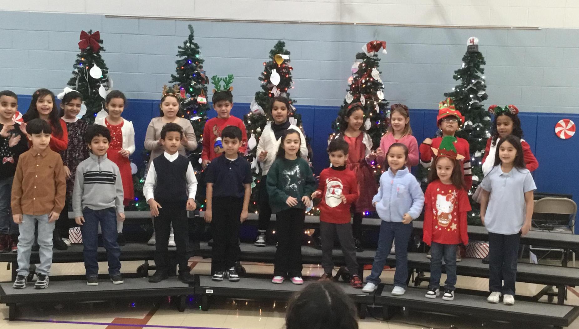 A group of children dressed in festive attire performing in front of decorated Christmas trees.