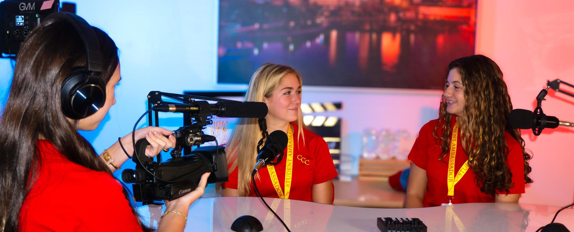 Two young ladies seated at a table look at one another with large microphones positioned near each, while a third girl wearing large headphones holds a large video camera pointed at them.