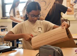 Girl with glasses cuts cardboard in a classroom setting.