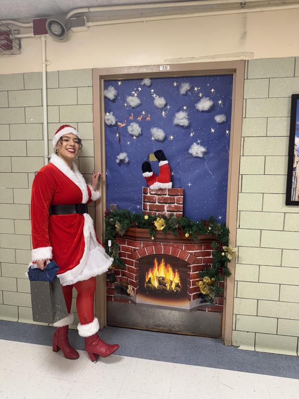 Ms. Rios beside her decorated door dressed in a Santa outfit. Door depicts a 3D creation of santa going down a chiminey with his legs poking out and a fireplace burning.