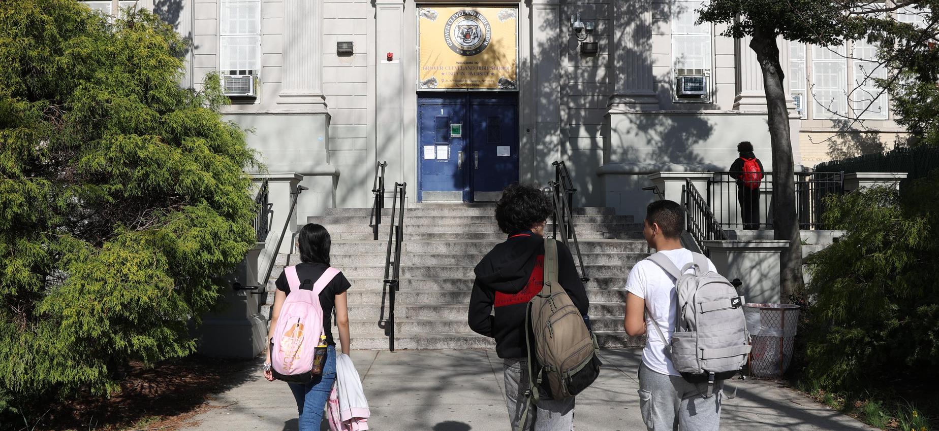 Three students approaching a school entrance with greenery around.