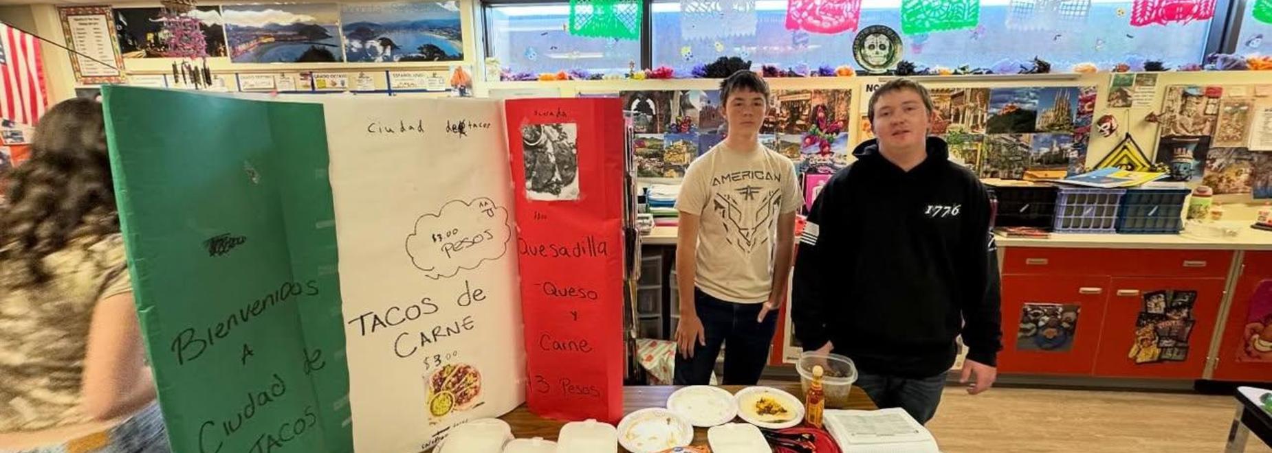 Two students standing in front of a project about Mexican food with decorations in a busy classroom.