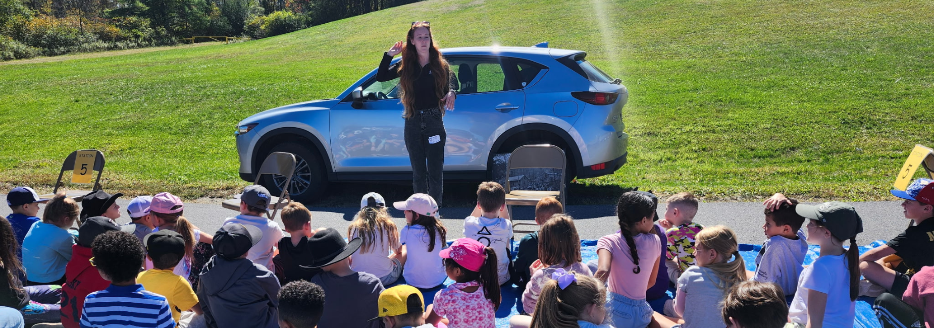 Children sitting on the ground listening to a presenter near a blue car.