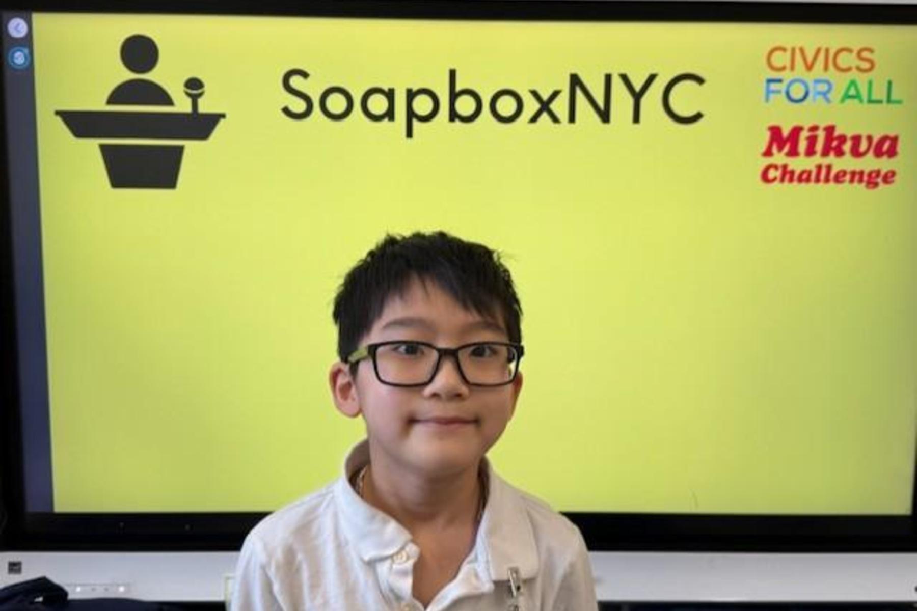 A child stands in front of a bright yellow backdrop with logos for SoapboxNYC and the Mikva Challenge.