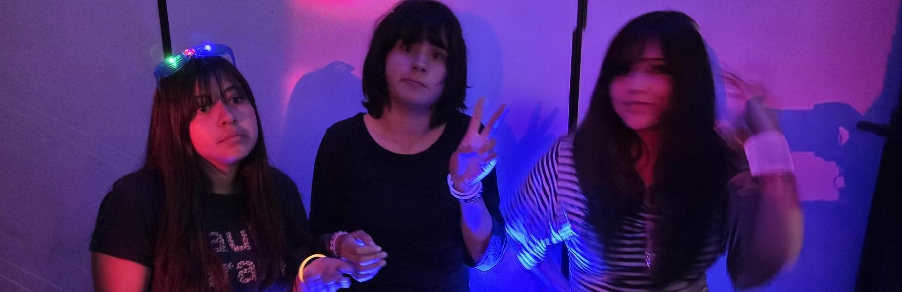 Three young women celebrate together in a dimly lit room with colorful lights.