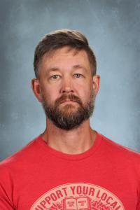 Man with a beard and short hair, wearing a red t-shirt, serious expression.