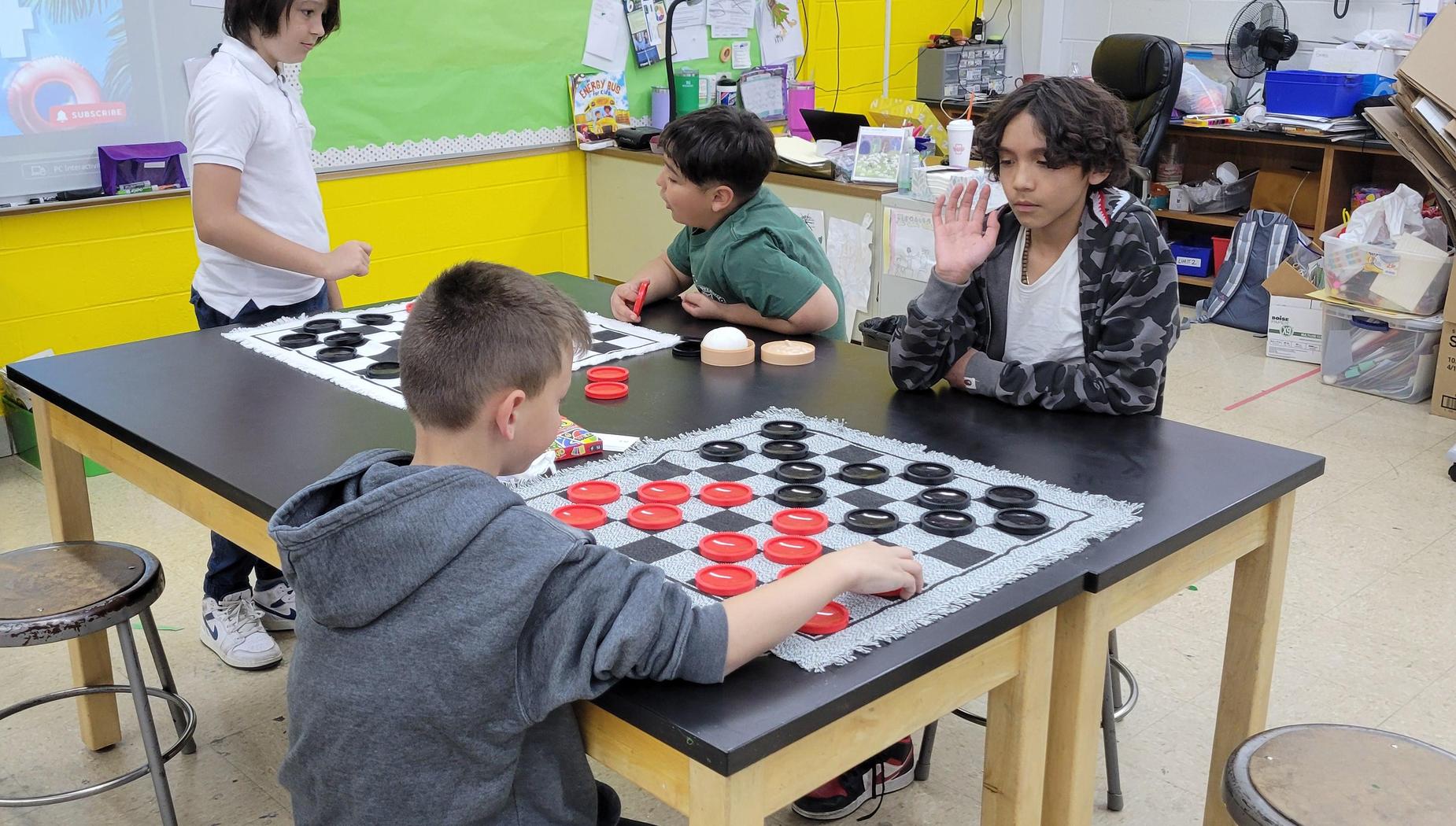 Children playing checkers at a classroom table with colorful game pieces.