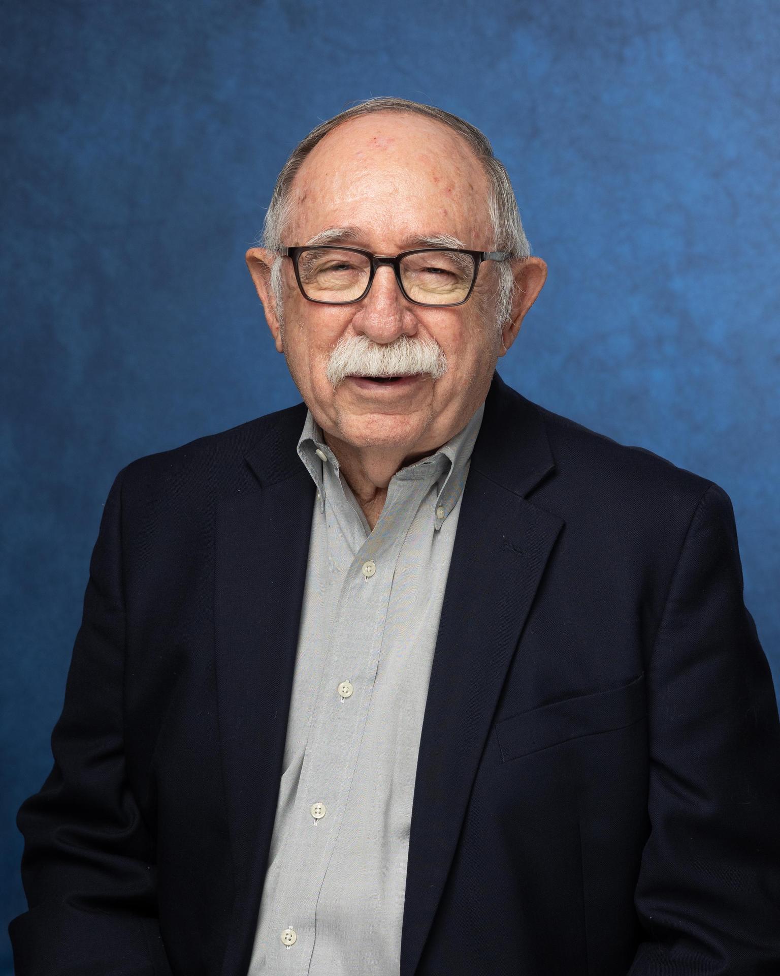 Smiling elderly man with glasses wearing a blazer against a blue background.
