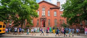 Children and teachers outside the red school building coming to school.