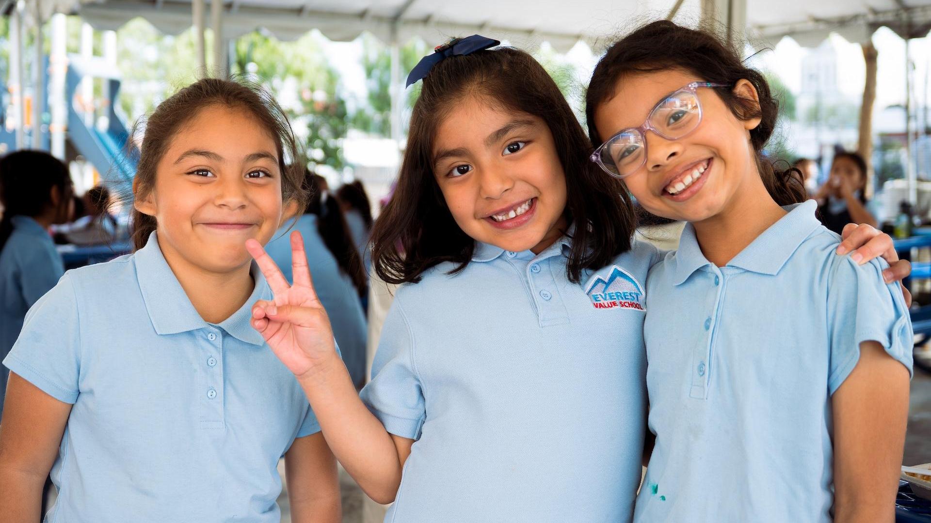 Three girls smiling and posing together in light blue shirts outdoors.