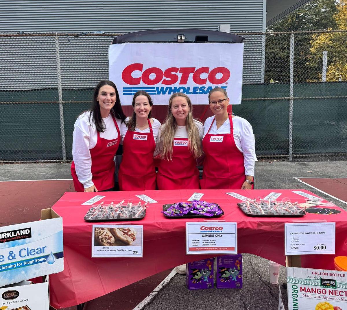 Four women in red aprons at a Costco booth with snacks and promotional materials.