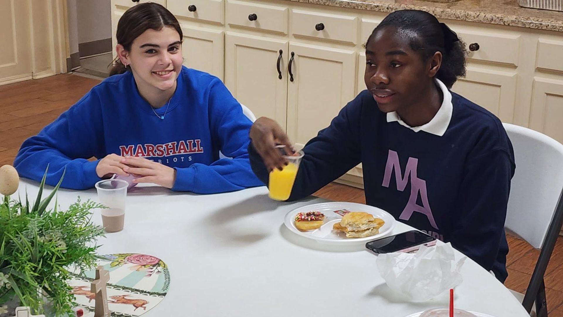 Two teenagers at a table with snacks and drinks, smiling at the camera.