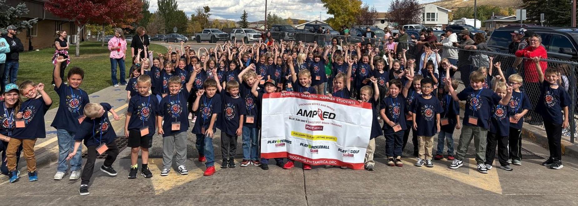 A large group of children wearing matching shirts, cheering with raised hands outdoors.