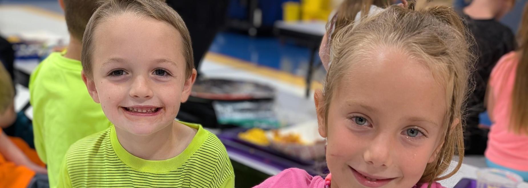 Two kids sitting together; one wears a pink shirt with cartoon dinosaurs.