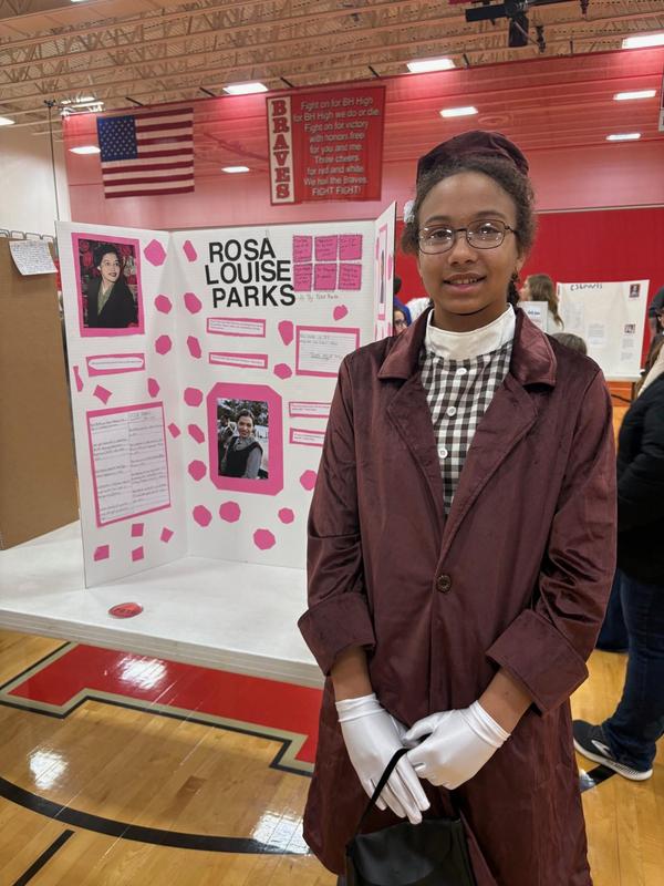 A student dressed as Rosa Parks stands beside her project board at a school event.