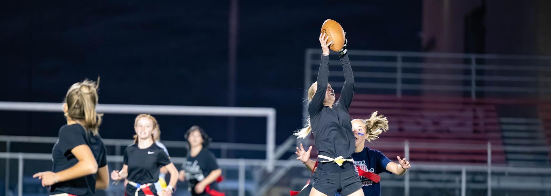 Girls playing flag football on a field at night.