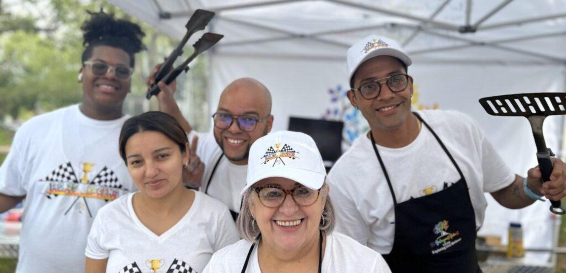 A diverse team of five smiling individuals at a grilling event, wearing matching shirts and hats.