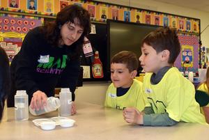 A woman demonstrates an experiment to two boys in yellow shirts at a classroom table.