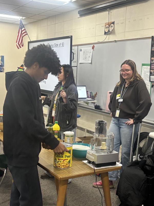 A student measures chickpeas while classmates assist with food preparation in class.