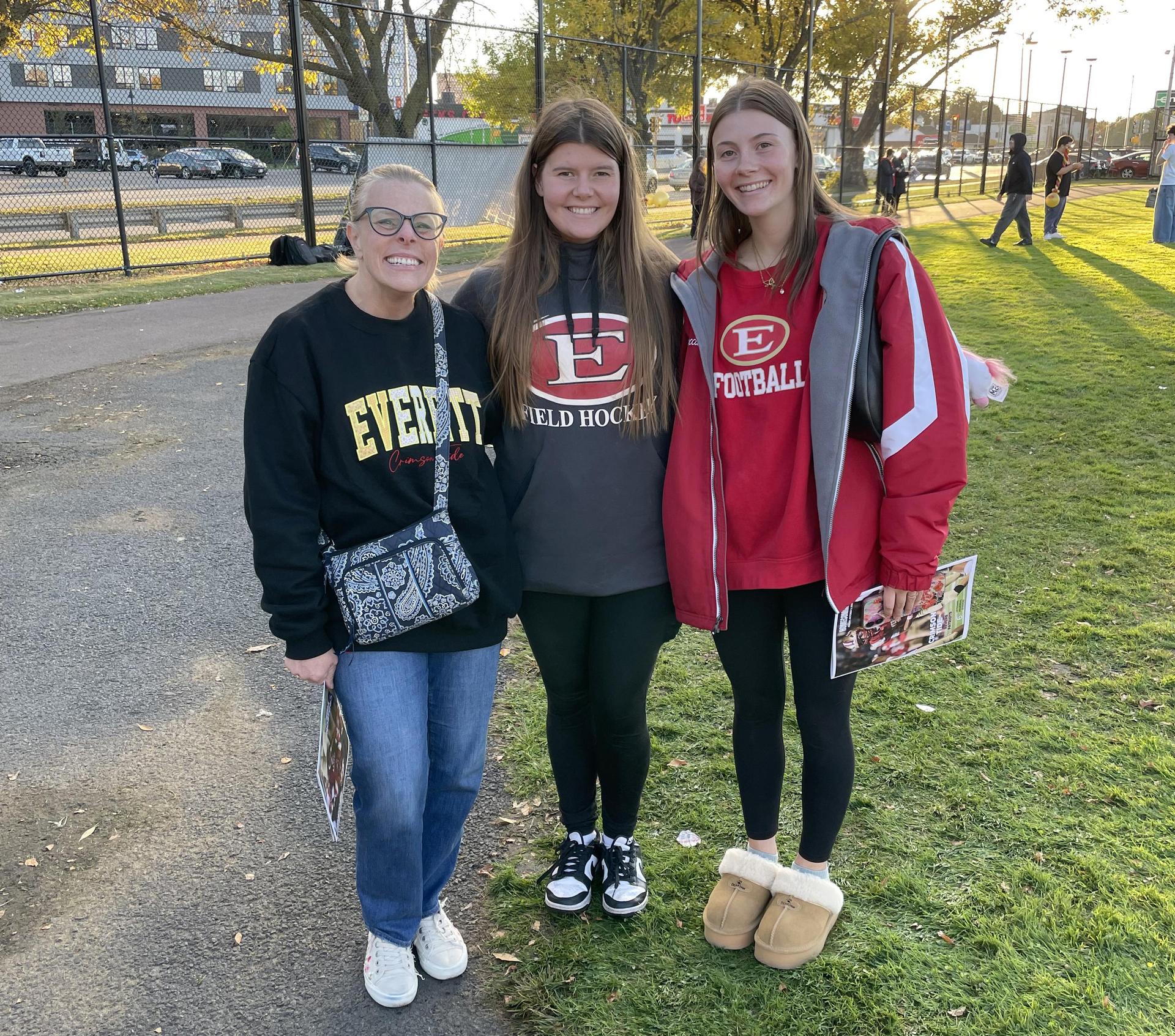 Three women posing together in a park during a football event.