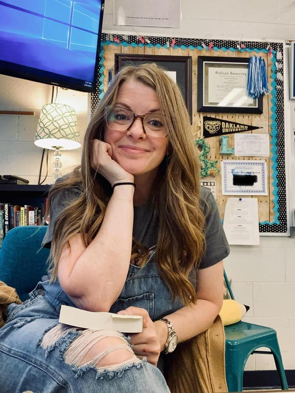 Female with long blond hair wearing glasses sitting down with a book in her hand.
