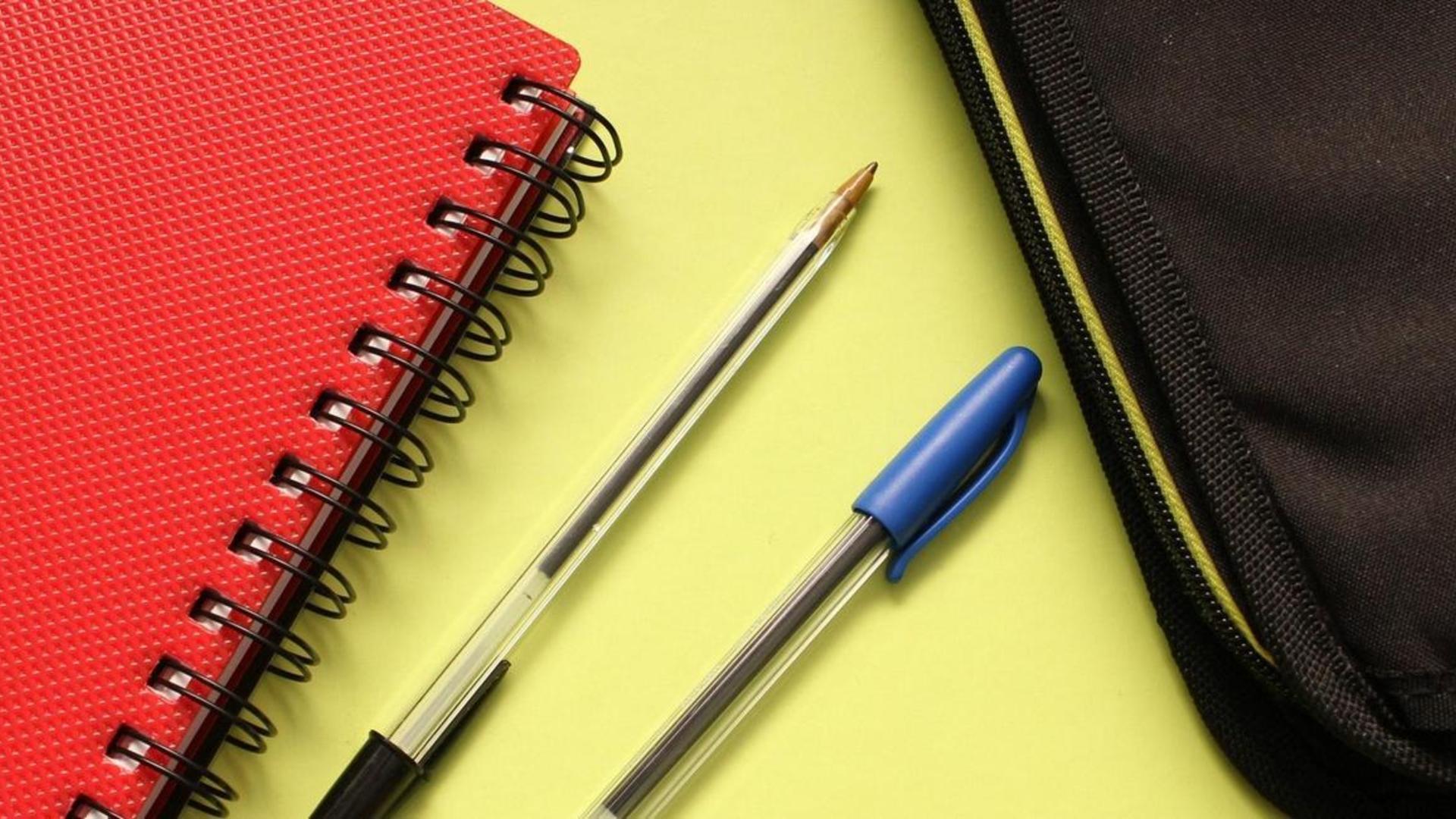 Red notebook, two pens, and a black pencil case on a yellow background.