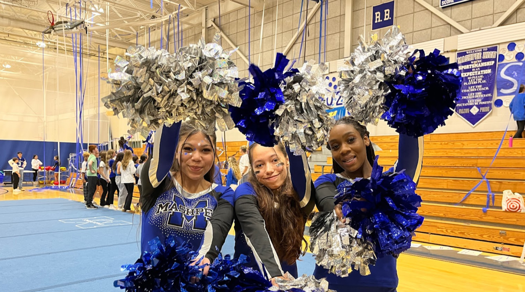 Three cheerleaders hold blue and silver pom-poms, smiling at the camera.