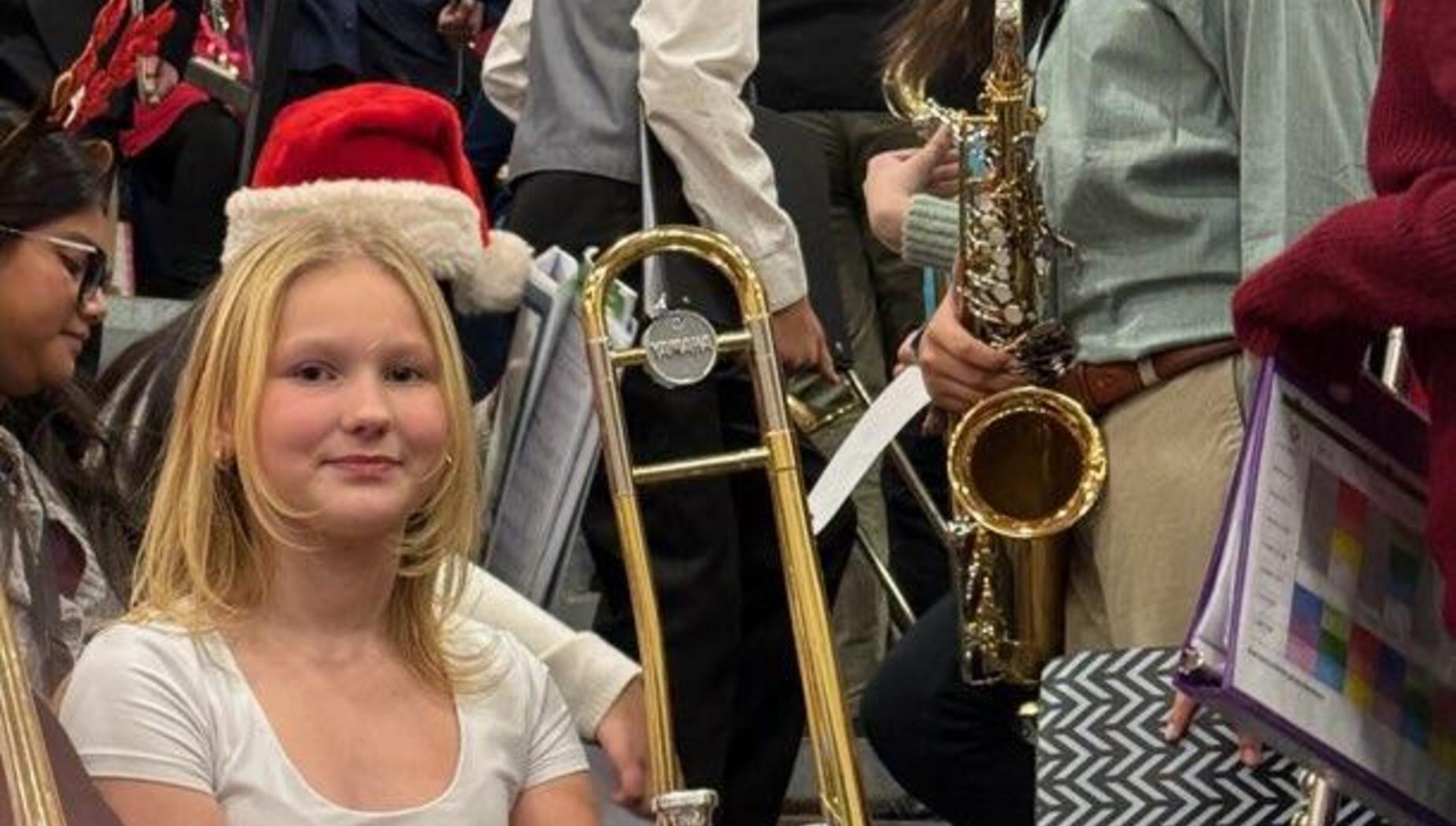 A girl with a trombone sitting in a crowded band scene with musicians in the background.
