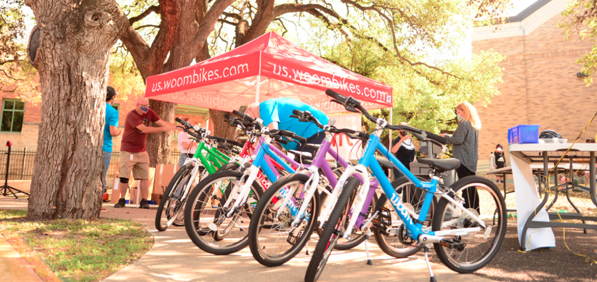 A display of colorful bikes presented to Texas School for the Deaf students by woom and 100BikesForKids. The event takes place under a red woom-branded tent, surrounded by trees and sunlight. Volunteers and staff are seen setting up and interacting with attendees, creating an atmosphere of excitement and community.