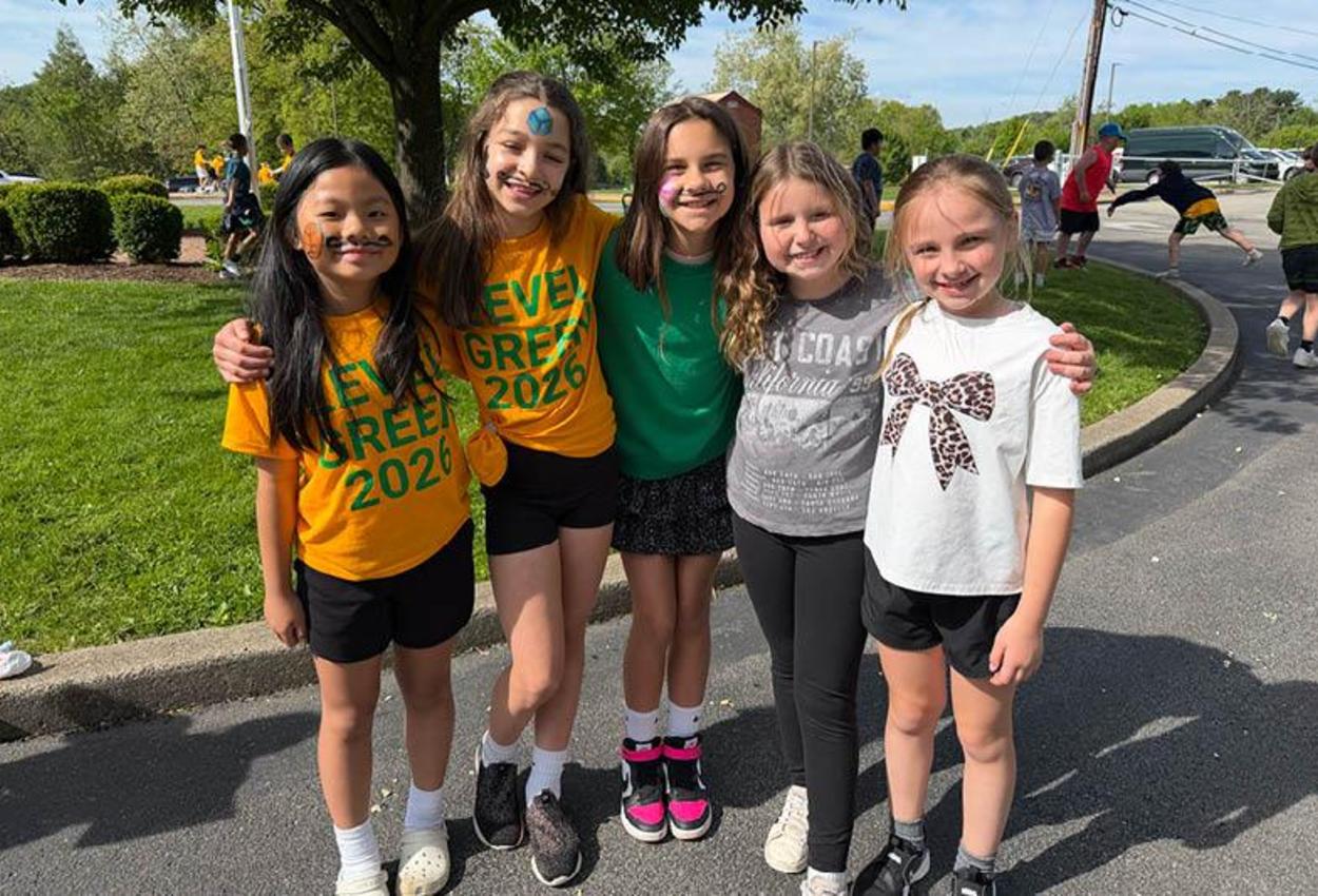 Five young girls in t-shirts stand arm in arm in a school parking lot, some with moustaches painted on their faces