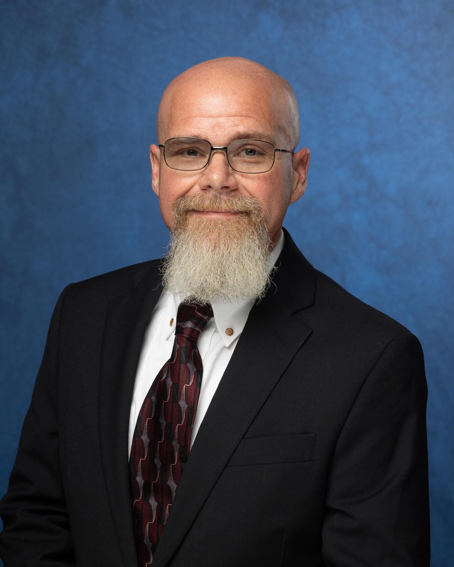 Bald man in glasses and tie wearing a black suit against a blue background.
