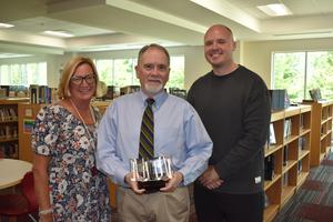 Three TJCA staff members pose in the Upper Campus library as the award recipient holds a crystal book-shaped plaque.