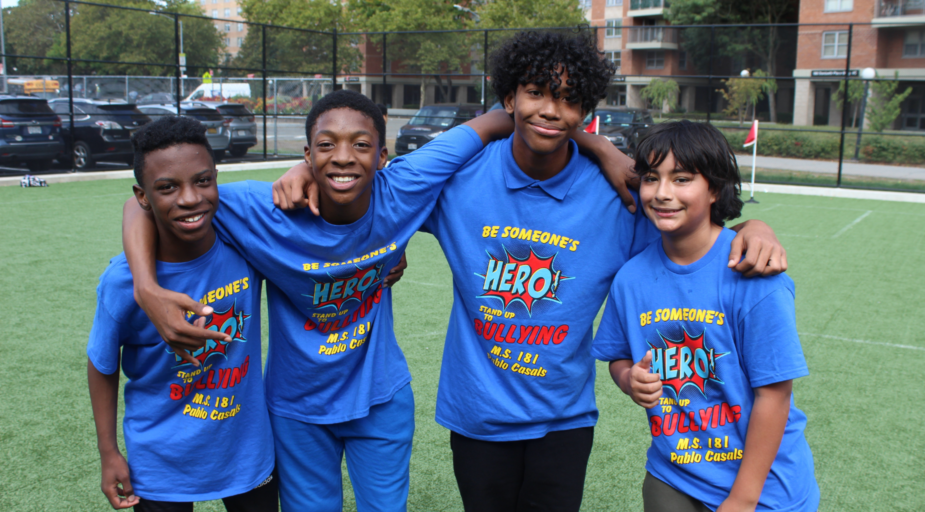 Four boys in blue shirts smiling together at a sports field.
