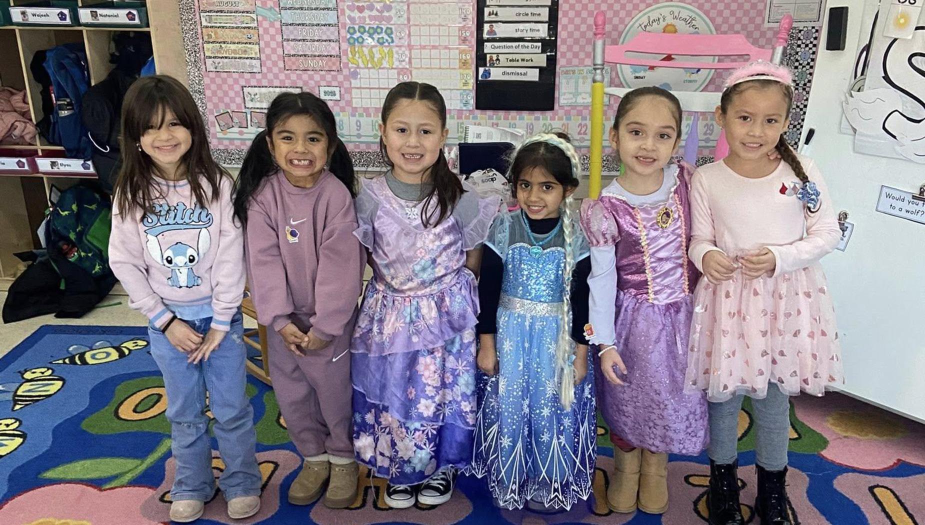 Group of girls dressed in colorful princess outfits standing on a patterned rug.