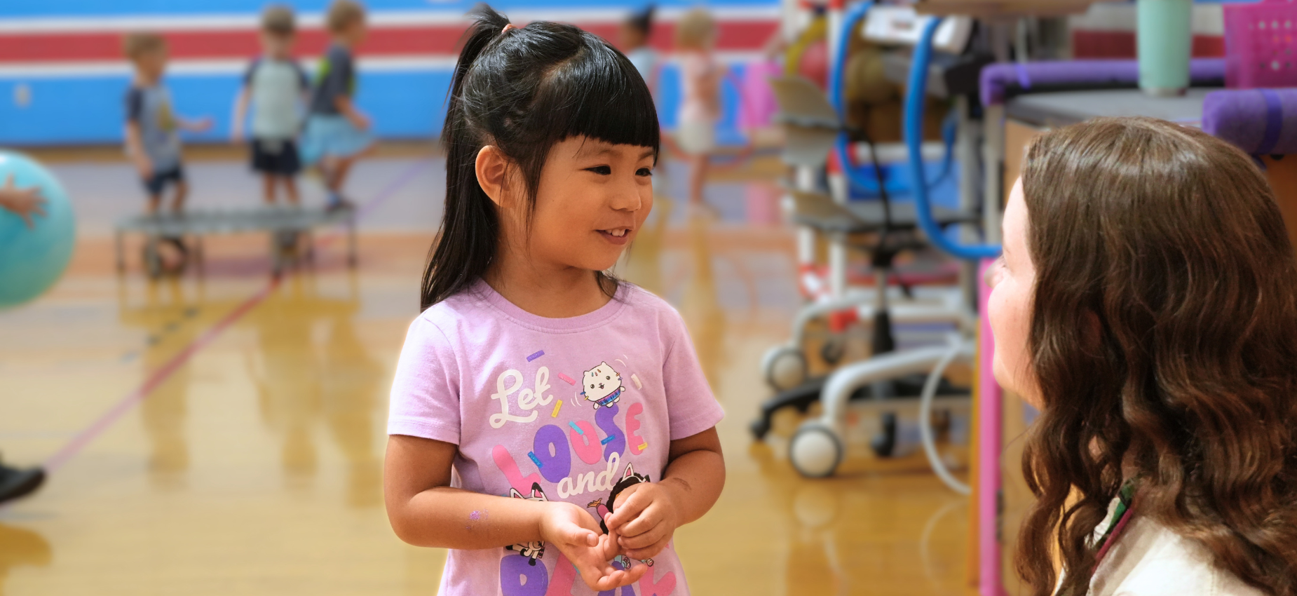 A young girl in a purple shirt smiles and talks to another person in a gym setting.